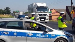 Na zdjęciu widać drogę, którą porusza się samochód ciężarowy oraz funkcjonariuszy Policji z Wydziału Ruchu Drogowego.