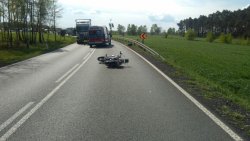Na zdjęciu widać przewrócony motocykl, który leży na środku drogi. Pojazd przewrócony w wyniku zdarzenia drogowego. W oddali widać ciąg pojazdów.