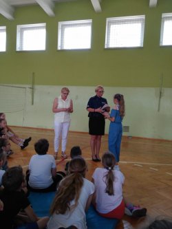 Na zdjęciu widać nauczycielkę ze Szkoły Podstawowej nr 1 w Ostrzeszowie, profilaktyka społecznego z Komendy Powiatowej Policji w Ostrzeszowie, uczniów siedzących twarzą do w/wymienionych osób oraz uczennicę, która otrzymuje nagrodę