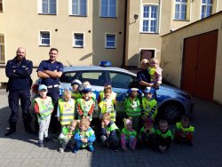 Na zdjęciu widać grupę przedszkolną, przy niej stoją funkcjonariusze Policji, w tle widac radiowóz oraz budynek KPP w Ostrzeszowie